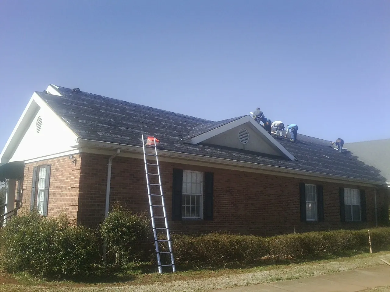 Crew tearing shingles off a brick ranch roof with a ladder set — Metal Roof Repair work by our crew in Ledbetter