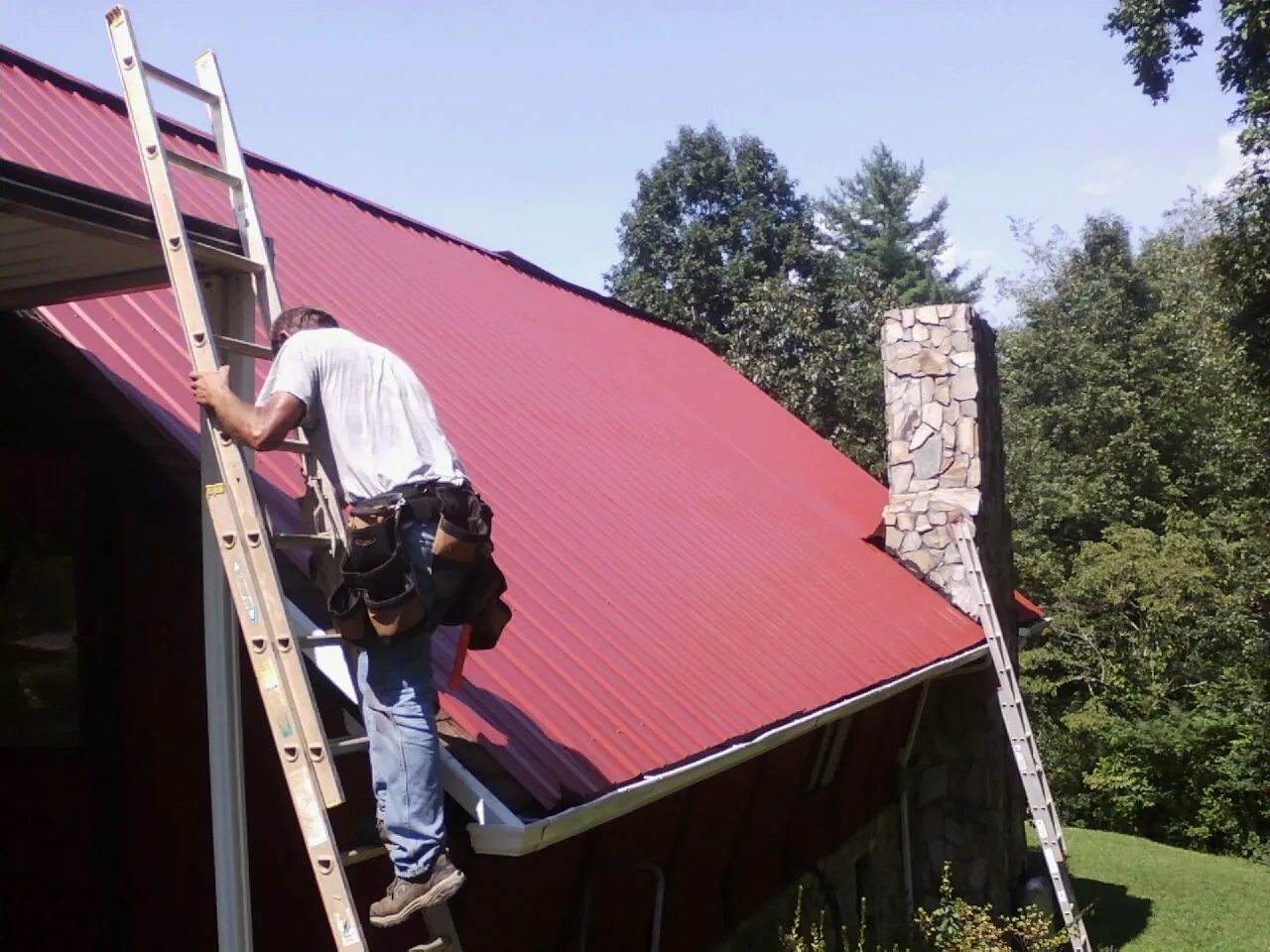 Roofer on a ladder beside a red metal roof and stone chimney — Soffit & Fascia Repair by Roofing Brothers in Ledbetter